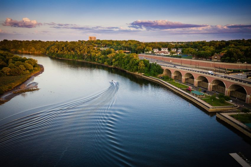 Aerial view of New Brunswick with a sunset over a boat speeding down the Raritan River next to a park and elevated highway near River Road Estates luxury home community